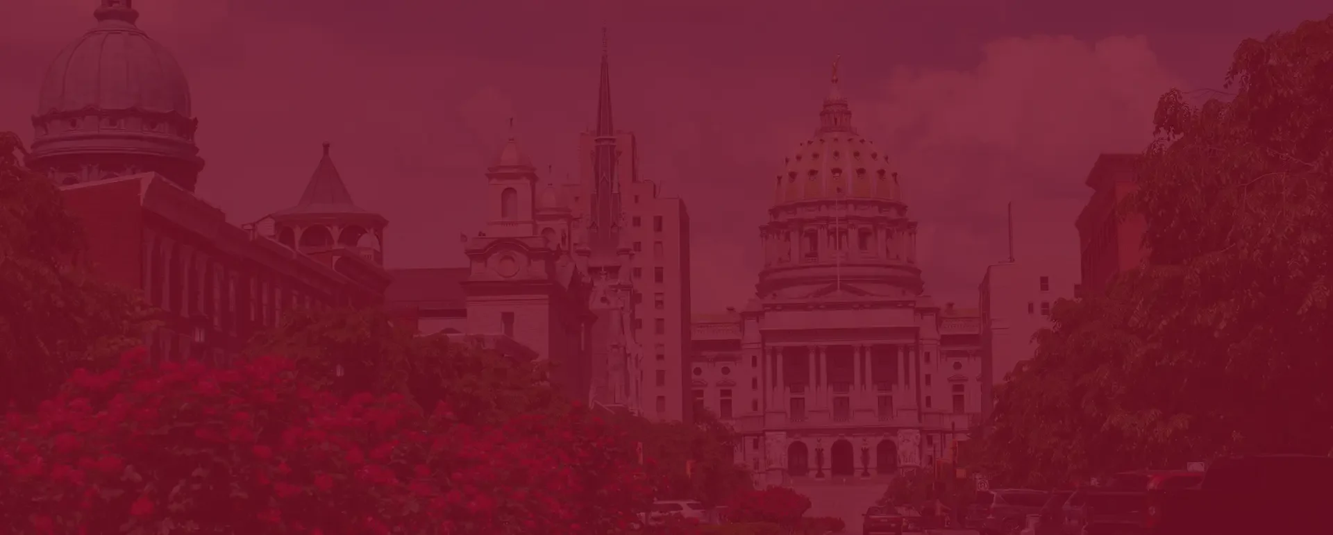 A wide shot of the Pennsylvania State Capitol in Harrisburg, with its green dome and classical facade, viewed from a street lined with vibrant red flowers in the foreground